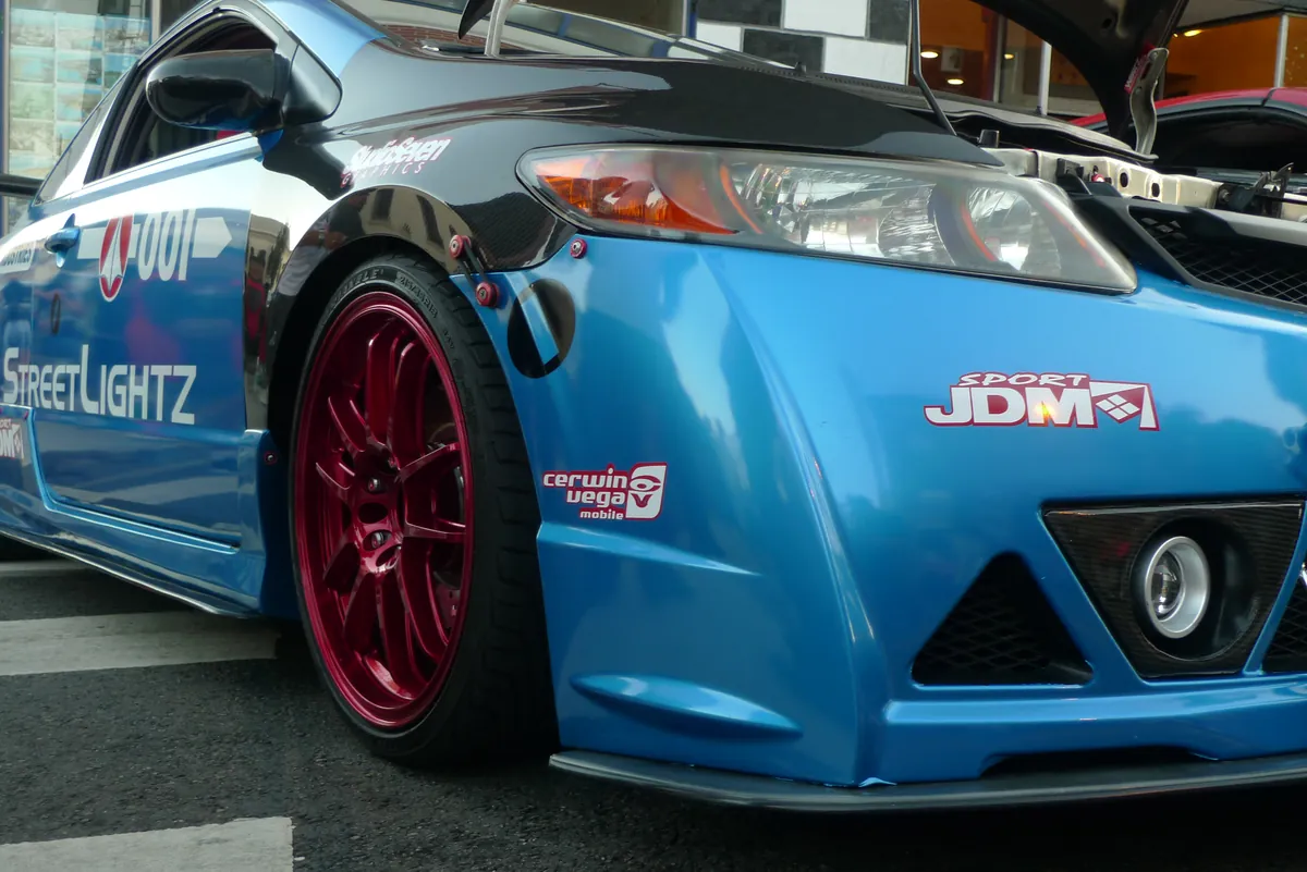 Front corner of a blue tuner car with red wheels at a car show