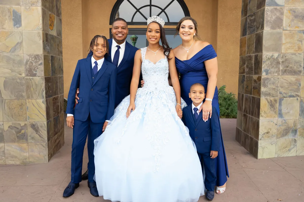 Family of five in navy formalwear gathered around a young woman in a pale blue ball gown and tiara