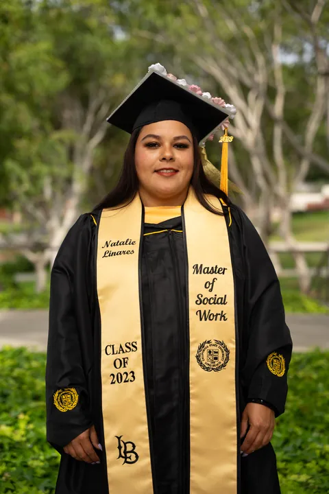 Graduate in a cap, gown, and gold stole standing among trees