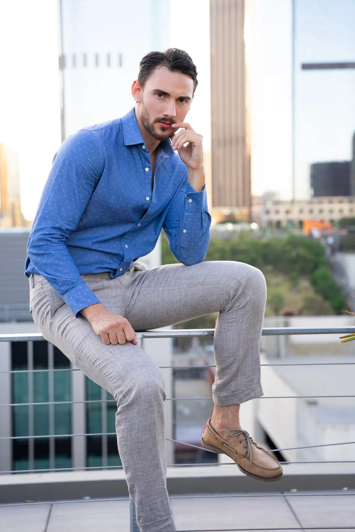 Man in a blue shirt seated on a rooftop railing with the downtown skyline behind him