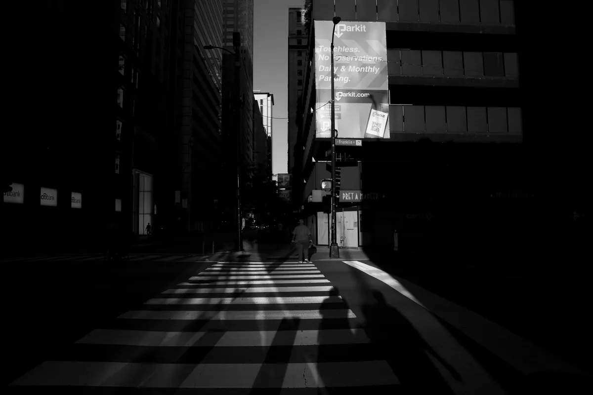 Black and white street photo of a lone pedestrian crossing a sunlit crosswalk between dark downtown buildings