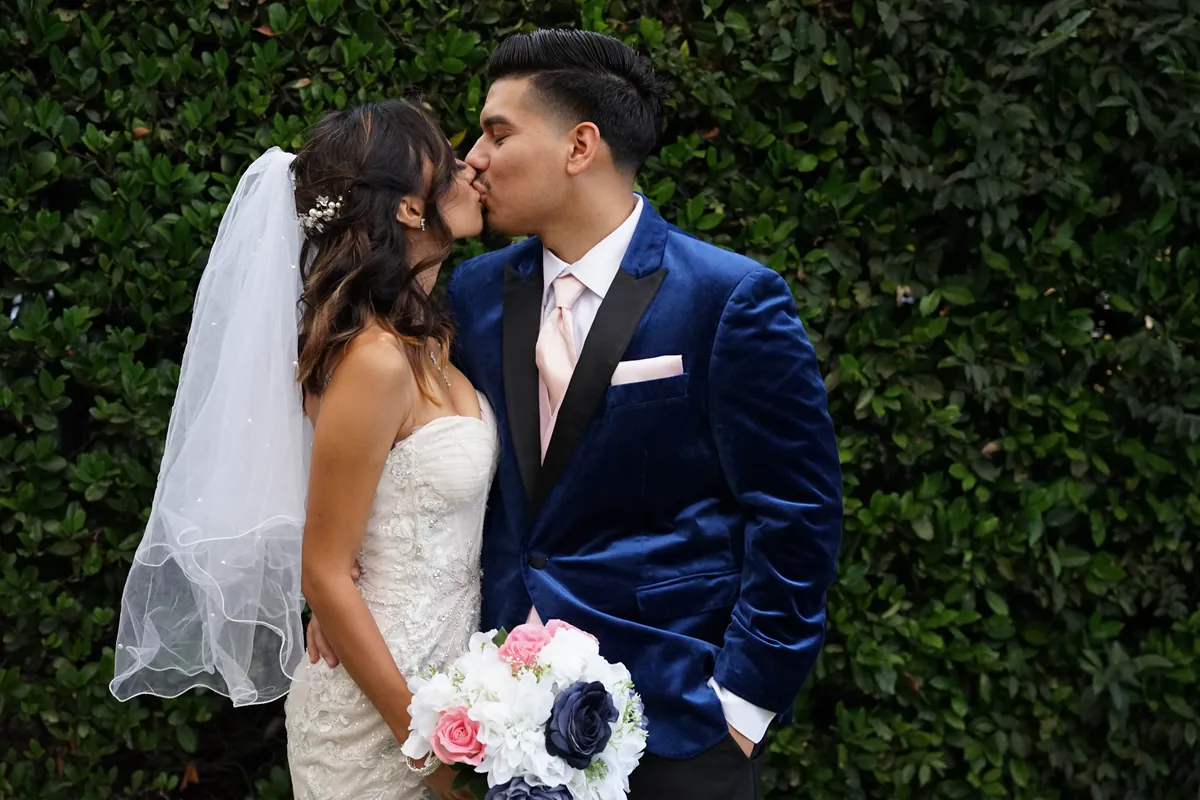 Bride and groom sharing a kiss in front of a tall green hedge